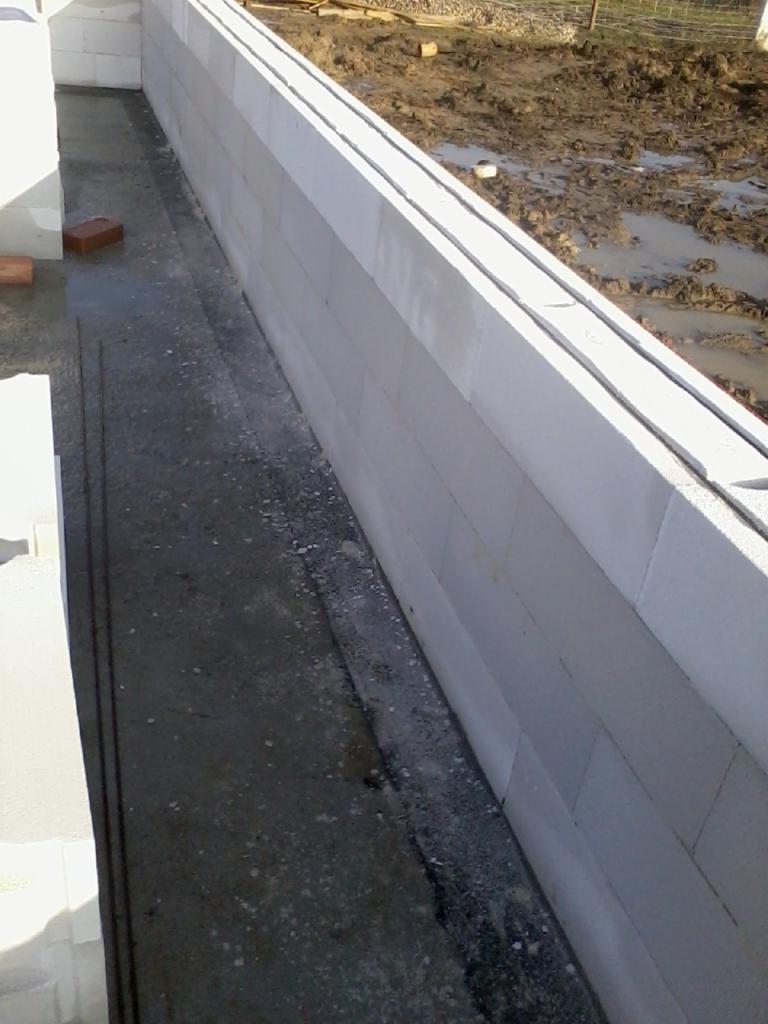 Ściana z betonu komórkowego w trakcie murowania, widoczna zaprawa i fragment zbrojenia. W tle błotniste podłoże.