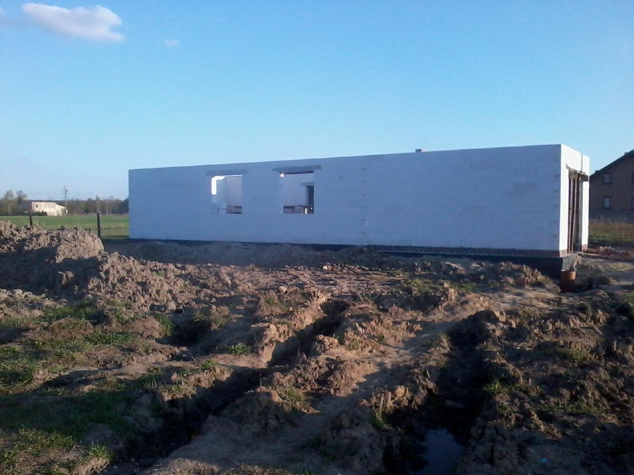 Niedokończony, parterowy dom z białego bloczka, widoczny na tle błękitnego nieba i zieleni. Wokół budynku ziemia z wykopami. Widoczne otwory okienne i drzwiowe.