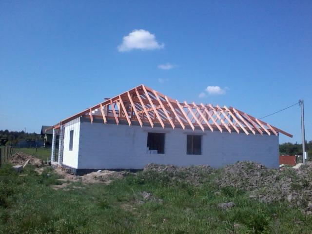 Wznoszony dom z białego bloczka, z częściowo wykonaną drewnianą konstrukcją dachu na tle błękitnego nieba i zielonej trawy. Widok z poziomu gruntu.