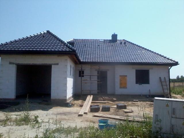 Niedokończony dom z ciemną dachówką, widoczne rusztowania i materiały budowlane. Stan surowy otwarty z garażem, w tle zielony krajobraz.