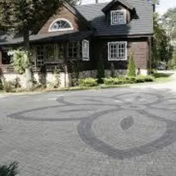 Ułożona kostka brukowa z dekoracyjnym wzorem kwiatowym na podjeździe przed domem z brązową elewacją i ciemnym dachem, otoczonym zielenią.