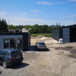 Teren składu opału w Jarosławiu z budynkiem biurowym, magazynem z otwartymi wrotami, stertami materiałów sypkich i workami z opałem. Na pierwszym planie zaparkowany samochód osobowy.