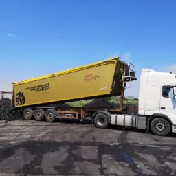 Żółta ciężarówka z naczepą wyładowuje czarny materiał sypki na betonowy plac pod błękitnym niebem.