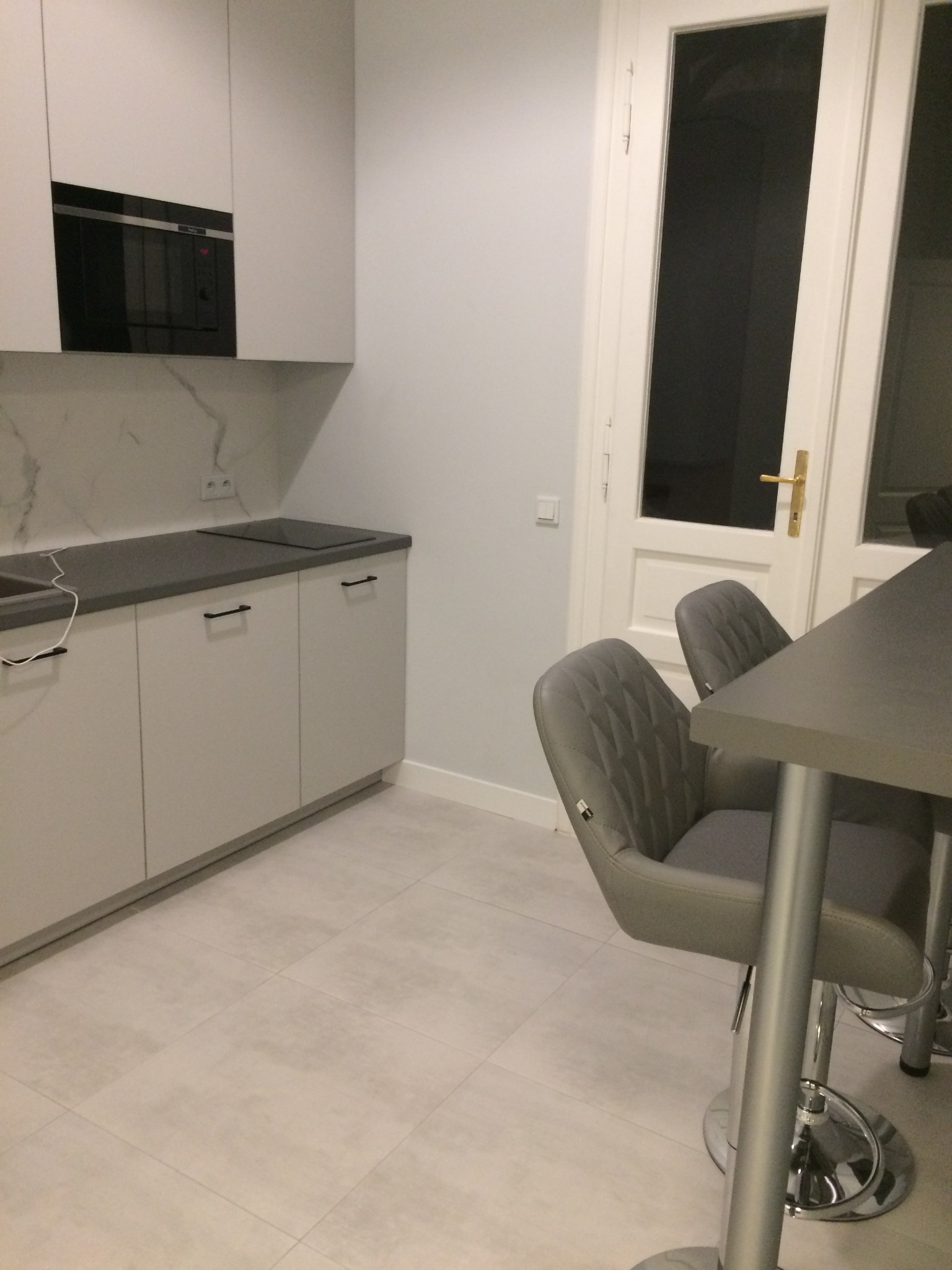 Jasna kuchnia w stylu skandynawskim z białymi szafkami, szarym blatem i marmurowym backsplashiem, uzupełniona dwoma szarymi krzesłami barowymi z pikowanymi oparciami i chromowanymi nogami.