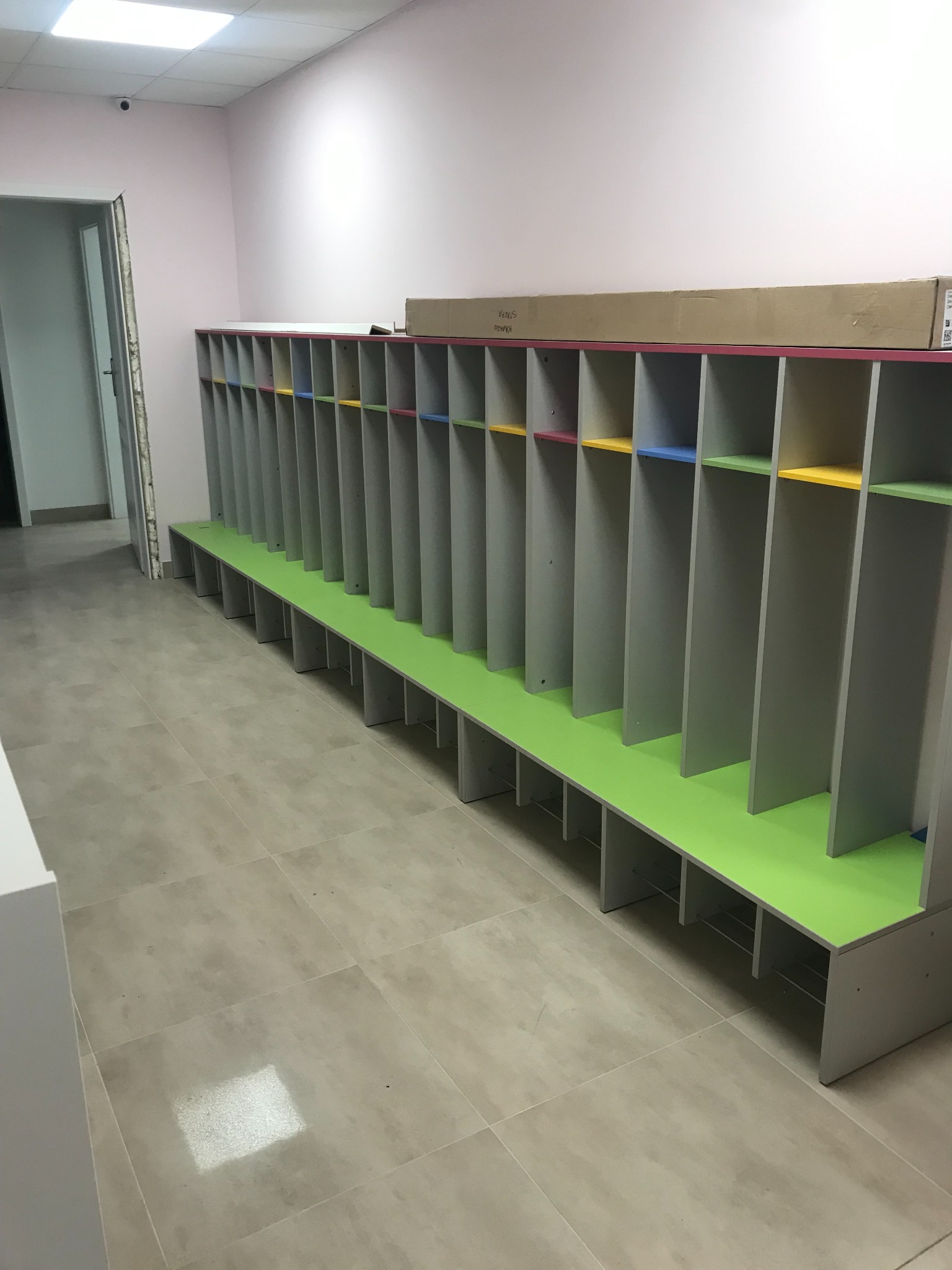 Długa szafka z przegródkami w przedszkolu lub szkole, pomalowana na szaro z kolorowymi akcentami wewnątrz przegródek (czerwony, żółty, niebieski, zielony) i zielonym siedziskiem, na szarych...