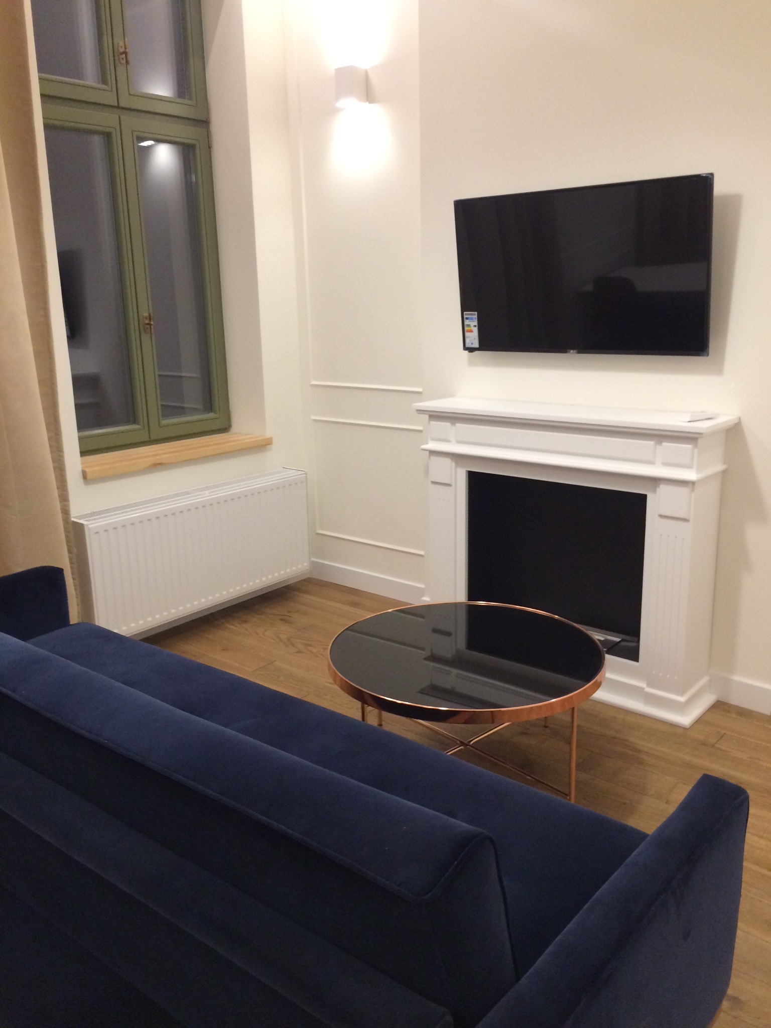 Salon z niebieską sofą, okrągłym stolikiem kawowym z miedzianymi nogami i kominkiem elektrycznym pod telewizorem, drewniana podłoga i okno z zieloną ramą.