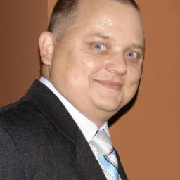 Portret mężczyzny w garniturze z krawatem w paski na tle jednolitej ściany. Uśmiechnięta twarz, jasne oczy. Profesjonalny wizerunek.