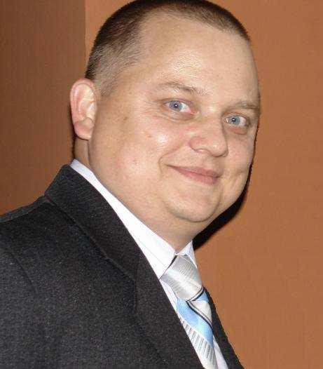 Portret mężczyzny w garniturze z krawatem w paski na tle jednolitej ściany. Uśmiechnięta twarz, jasne oczy. Profesjonalny wizerunek.