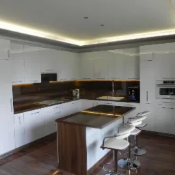 Błyszcząca, nowoczesna kuchnia z białymi frontami, brązowym blatem i wyspą. Oświetlenie LED pod szafkami górnymi i nad wyspą. Cztery białe hokery przy wyspie.