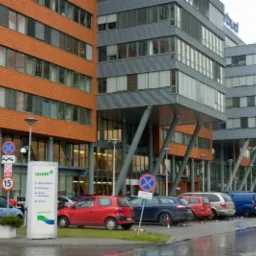 Nowoczesny budynek biurowy z pomarańczową i szarą fasadą, logo 'rbud' na szczycie. Na pierwszym planie znak Luxmed i zaparkowane samochody.