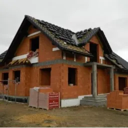 Dom murowany w trakcie budowy, widoczne bloczki Porotherm, częściowo położony dach z czarnej dachówki. Stan surowy otwarty. Na pierwszym planie palety z materiałami.