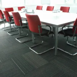 Wykładzina dywanowa w biurze z wzorem w kwadraty, ciemnoszara z czerwonymi i białymi akcentami, widoczne krzesła biurowe z czerwonym oparciem i biały stół konferencyjny.