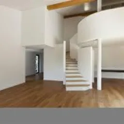 Jasne, przestronne wnętrze domu z drewnianą podłogą, spiralnymi schodami i widoczną kuchnią w tle. Naturalne światło podkreśla fakturę drewna.