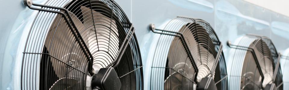 Rząd trzech wentylatorów przemysłowych z czarnymi osłonami, zamontowanych na jasnoniebieskiej ścianie, widocznych z boku pod kątem, tworzących powtarzalny wzór.