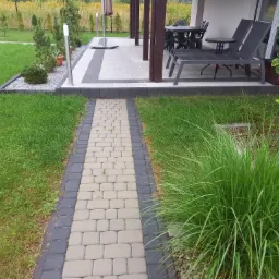 Ułożona z kostki brukowej ścieżka w ogrodzie prowadząca do tarasu z meblami ogrodowymi i leżakami.