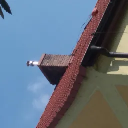 Fragment dachu z czerwoną dachówką, rynną i kominem wentylacyjnym w słoneczny dzień.