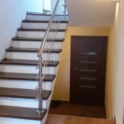 Wykończone schody z drewnianymi stopniami i metalową balustradą w jasnym wnętrzu. Widoczna brązowa stolarka drzwiowa i fragment podłogi z płytek.