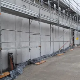 Ściana budynku w trakcie prac tynkarskich, z widocznymi rusztowaniami i zabezpieczeniami. Szara, surowa powierzchnia tynku w tle.