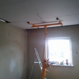 Montaż płyty gipsowej na suficie za pomocą podnośnika. Widoczne narzędzia i częściowo wykończony sufit w pomieszczeniu z oknem i drabiną.