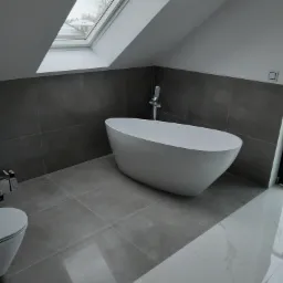 Nowoczesna łazienka na poddaszu z wolnostojącą białą wanną, szarymi płytkami ściennymi i podłogowymi oraz oknem dachowym z widokiem na zaśnieżone dachy.