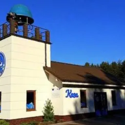 Nietypowy budynek kasy biletowej z elementami latarni morskiej i niebieską kopułą na dachu. Na ścianie logo z motywem morskim. W tle las iglasty, błękitne niebo.