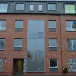 Fasada budynku z czerwonej cegły z nowoczesnymi oknami i szklaną ścianą. Nietypowe wzory dekoracyjne po bokach. Widoczny fragment czerwonego pojazdu.