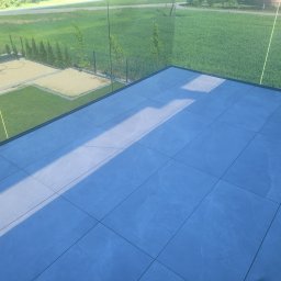 SZYMON GOLA BUDOWNICTWO - Nowoczesny taras z szarych płytek ceramicznych, otoczony szklaną balustradą z widokiem na zielony ogród. Minimalistyczny design i naturalne światło.