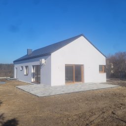 SZYMON GOLA BUDOWNICTWO - Nowoczesny, parterowy dom z białymi ścianami i szarą dachówką, z dużymi przesuwnymi oknami i wybrukowanym tarasem, na tle błękitnego nieba i drzew bez liści.