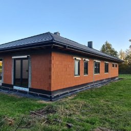SZYMON GOLA BUDOWNICTWO - Niski dom z czerwonej cegły z czarnym dachem, widok z boku na tle zielonego trawnika i błękitnego nieba. Widoczne okna i drzwi w czarnych ramach.