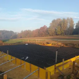 Zbrojenie płyty fundamentowej w żółtym szalunku, widok z góry, na tle jesiennego krajobrazu z drzewami o różnobarwnych liściach i błękitnym niebem.