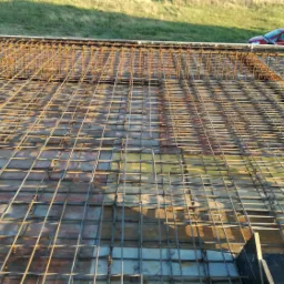 Siatka zbrojeniowa na szalunku, przygotowana do zalania betonem, z widocznym czerwonym samochodem w tle.