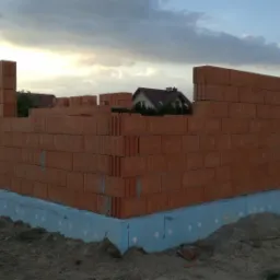 Wznoszone ściany z czerwonej cegły na fundamencie ocieplonym niebieskim styropianem, widok na budowę domu jednorodzinnego w trakcie realizacji, pochmurne niebo w tle.