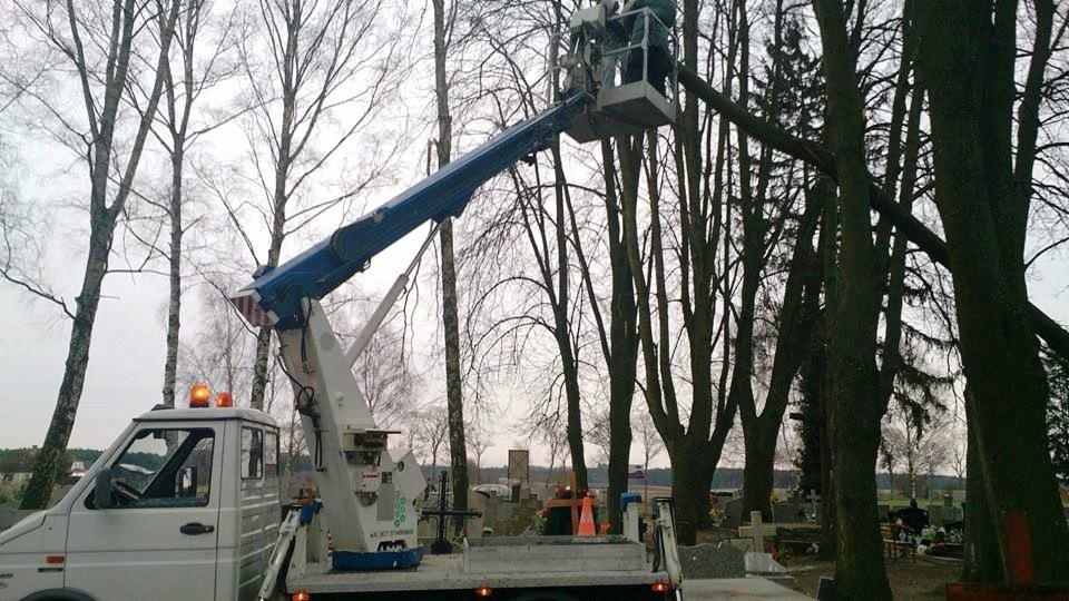 Wysięgnik na cmentarzu podczas przycinania drzew; widoczna platforma robocza z osobą, biała ciężarówka z pomarańczowymi światłami ostrzegawczymi i liczne nagrobki w tle.