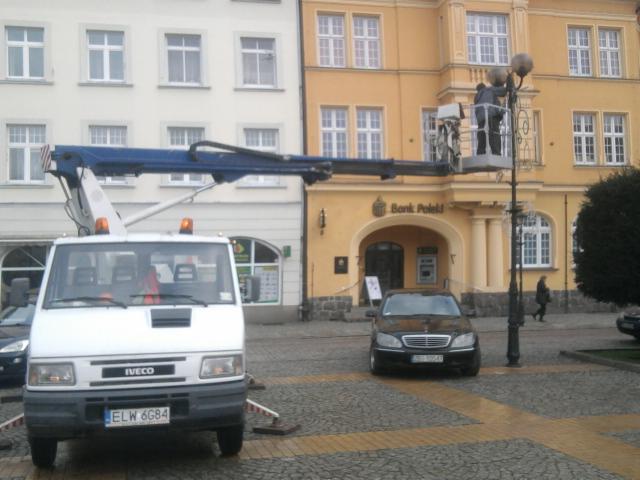 Pracownik na podnośniku koszowym naprawia latarnię na placu miejskim. W tle budynki, w tym Bank Polski. Na pierwszym planie samochód dostawczy Iveco.