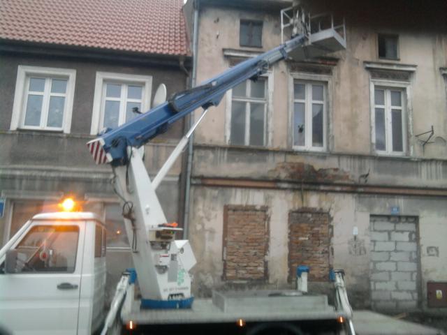 Podnośnik koszowy na tle starej kamienicy. Platforma robocza uniesiona, gotowa do prac na wysokości. Widoczne ceglane zamurowania okien i uszkodzony tynk.