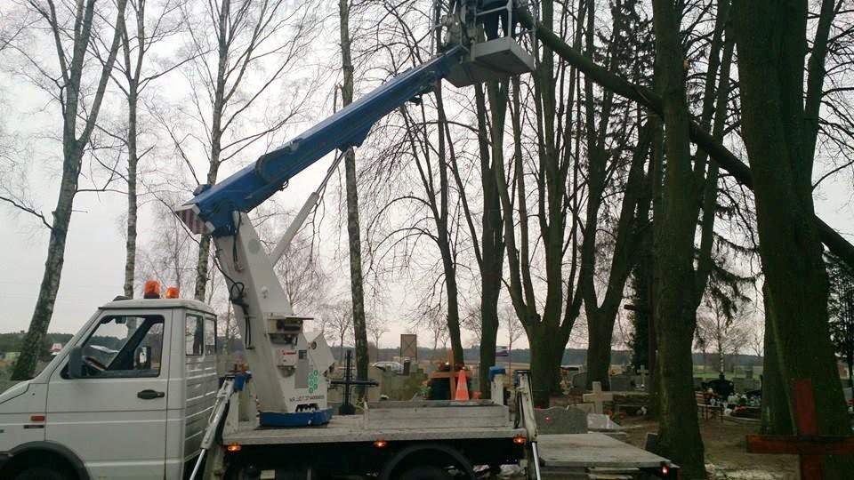 Podnośnik koszowy przy drzewach na cmentarzu, przygotowany do wycinki gałęzi. Widoczny fragment nagrobków i krzyży. Pochmurny dzień.