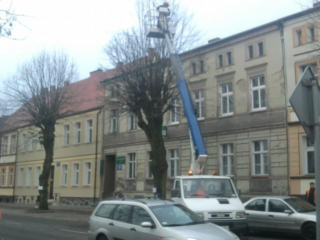 Pracownik na podnośniku koszowym przycina drzewo w miejskiej zabudowie. Biały samochód dostawczy i zaparkowane auta w tle. Jesienna aura.