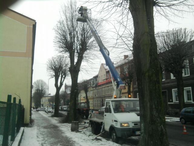 Podnośnik koszowy przy ulicy, używany do przycinania drzew w zimowy dzień. Na chodniku leży śnieg, w tle budynki mieszkalne.