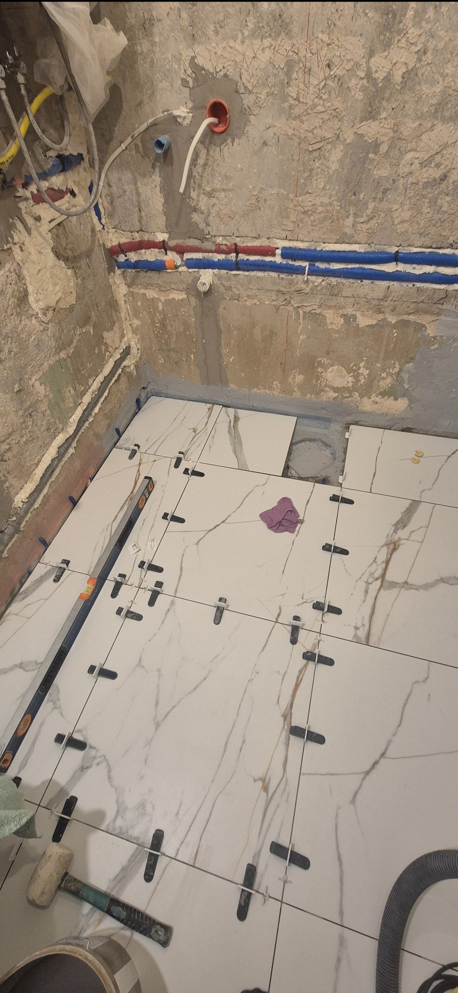 Układanie białych płytek imitujących marmur w łazience. Widoczne poziomica, krzyżyki dystansowe, fragment ściany z instalacją wodną i kanalizacyjną.