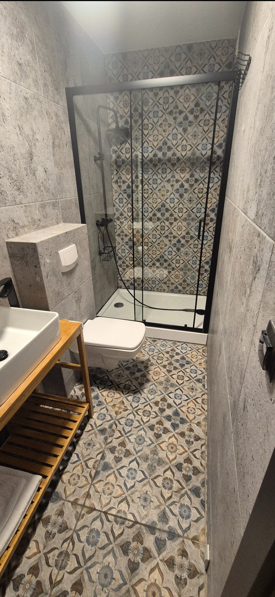 Wykończona łazienka z kabiną prysznicową typu walk-in, umywalką na drewnianym stelażu i toaletą. Na podłodze i ścianach wzorzyste płytki w odcieniach szarości i beżu.
