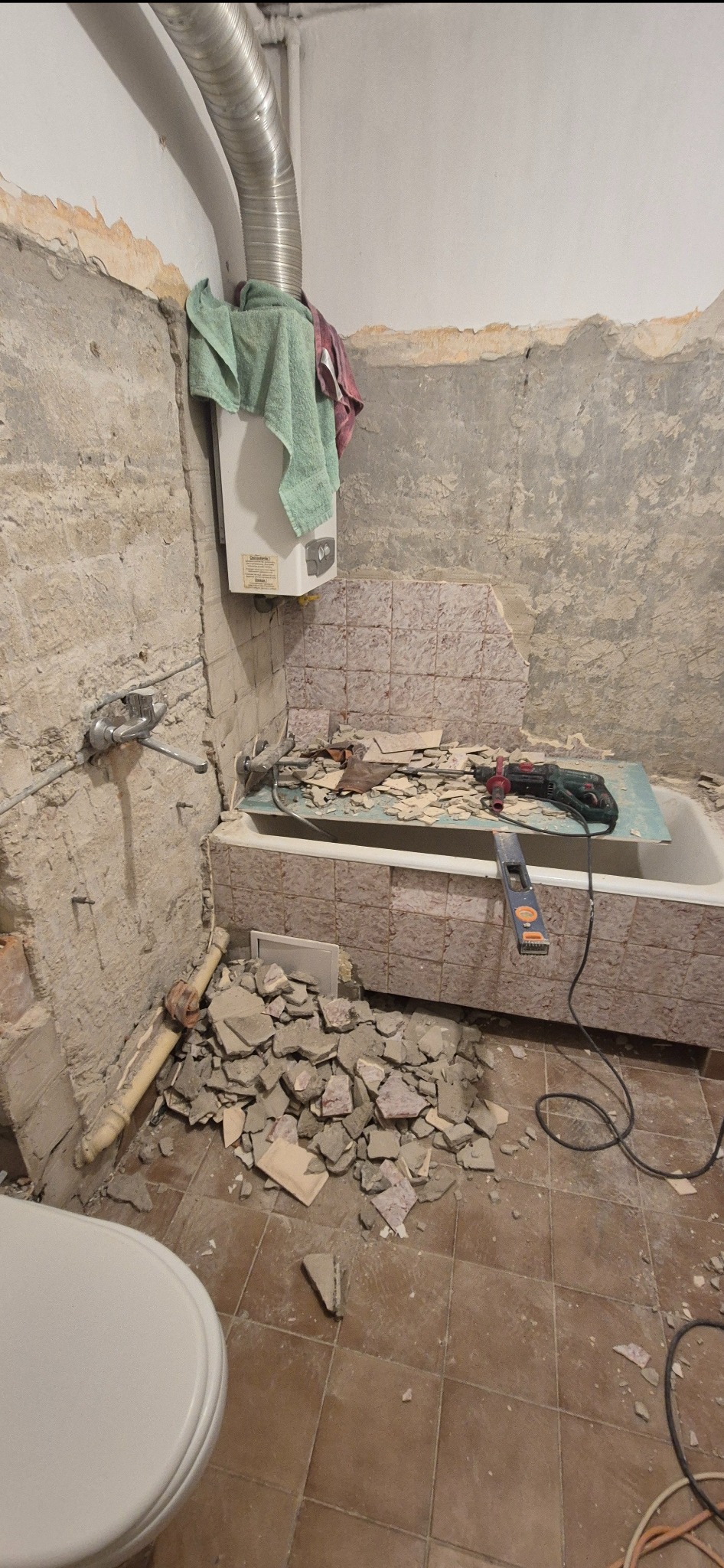 Demontaż płytek w łazience: gruz na podłodze, wanna z resztkami płytek, ściany częściowo bez okładziny. Widoczny bojler i narzędzia.