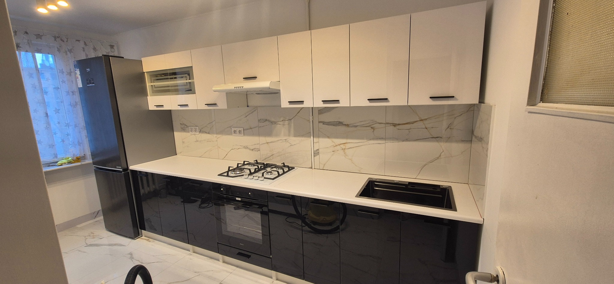 Nowoczesna kuchnia z białymi szafkami górnymi i czarnymi dolnymi, biały blat, marmurowe płytki na ścianie, srebrna lodówka. Minimalistyczny design.