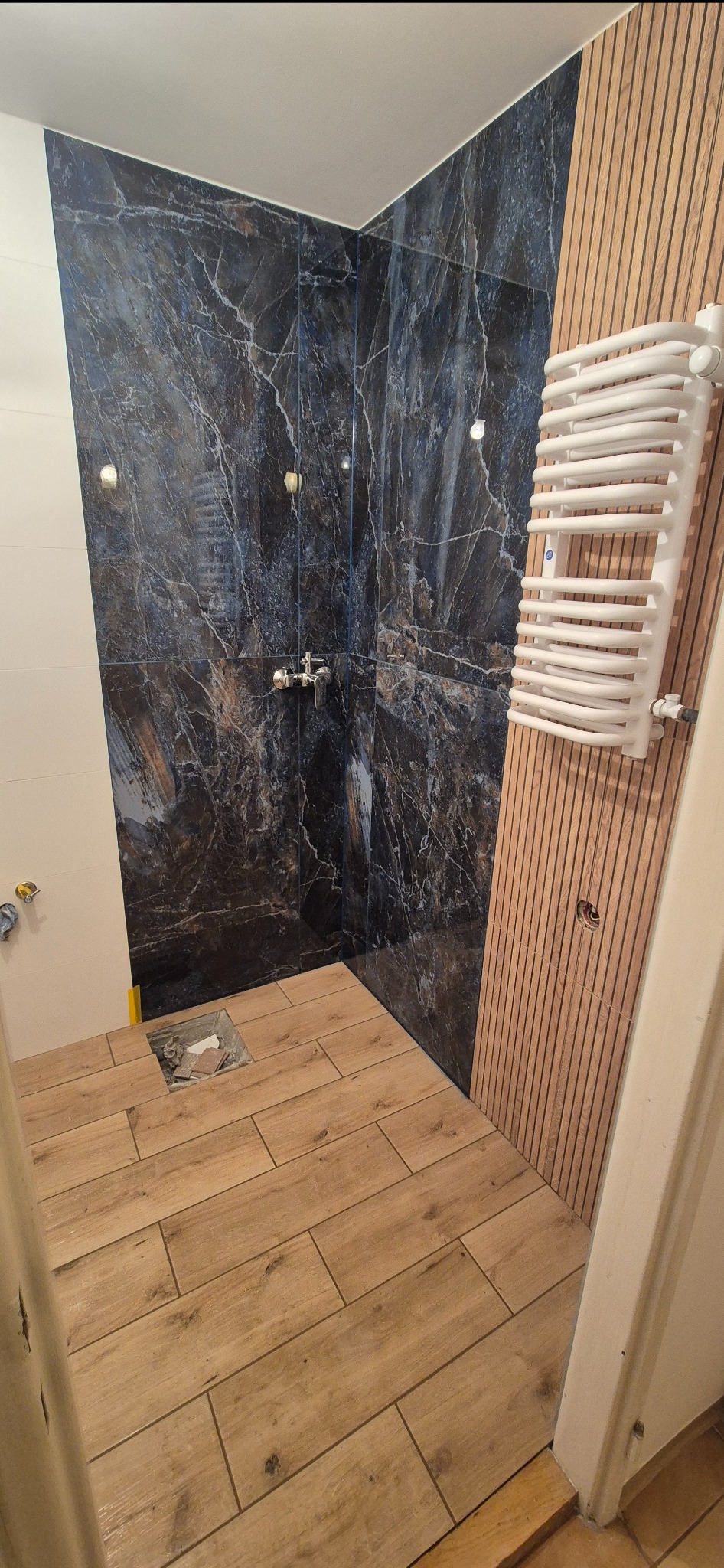 Wykończona łazienka z drewnopodobną podłogą, ciemnymi płytkami imitującymi marmur pod prysznicem i grzejnikiem drabinkowym na drewnianej ściance.