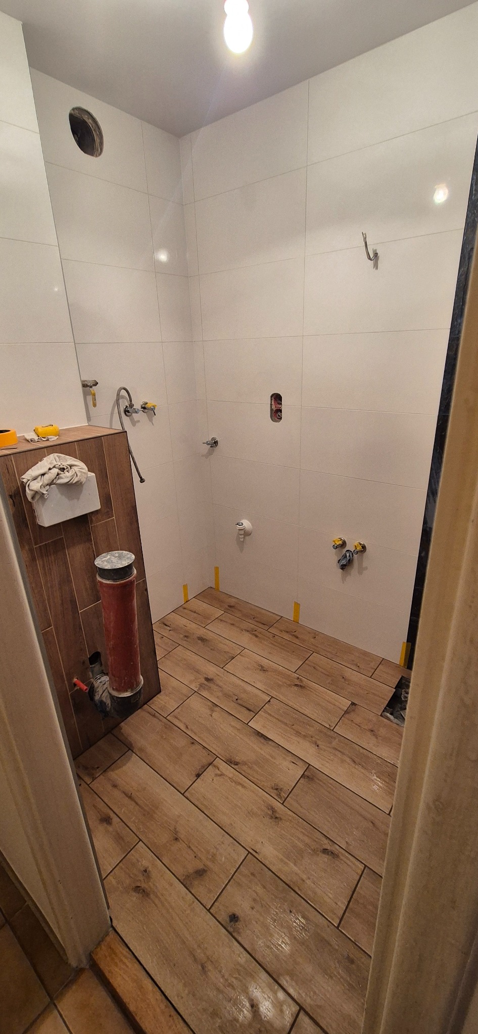 Łazienka w trakcie wykończenia: ułożona drewnopodobna podłoga, białe płytki na ścianach, widoczne instalacje wodne i kanalizacyjne, przygotowania do montażu toalety.