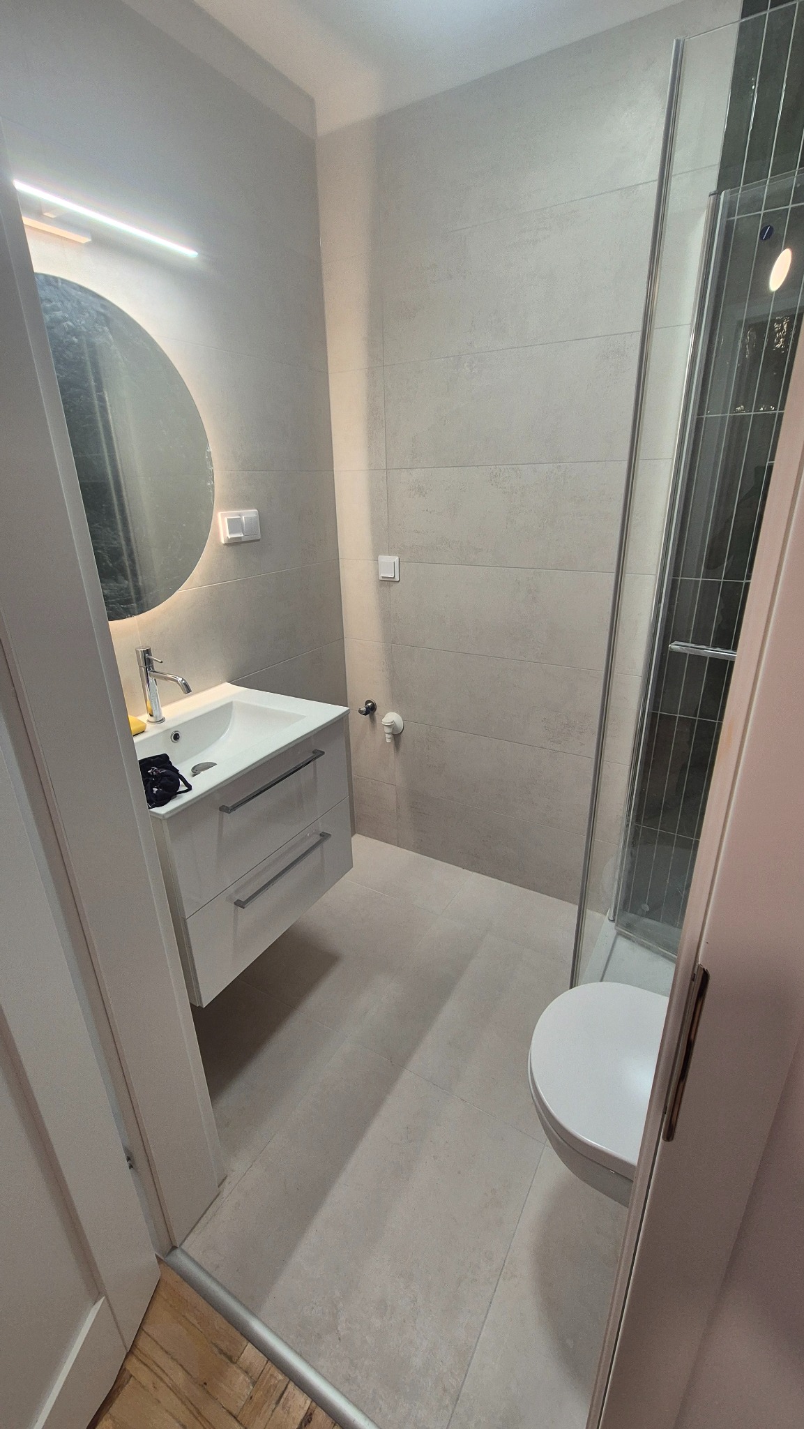 Jasna łazienka z kabiną prysznicową, umywalką z szafką i okrągłym lustrem z podświetleniem LED. Minimalistyczny design, szare płytki, widok zza otwartych drzwi.