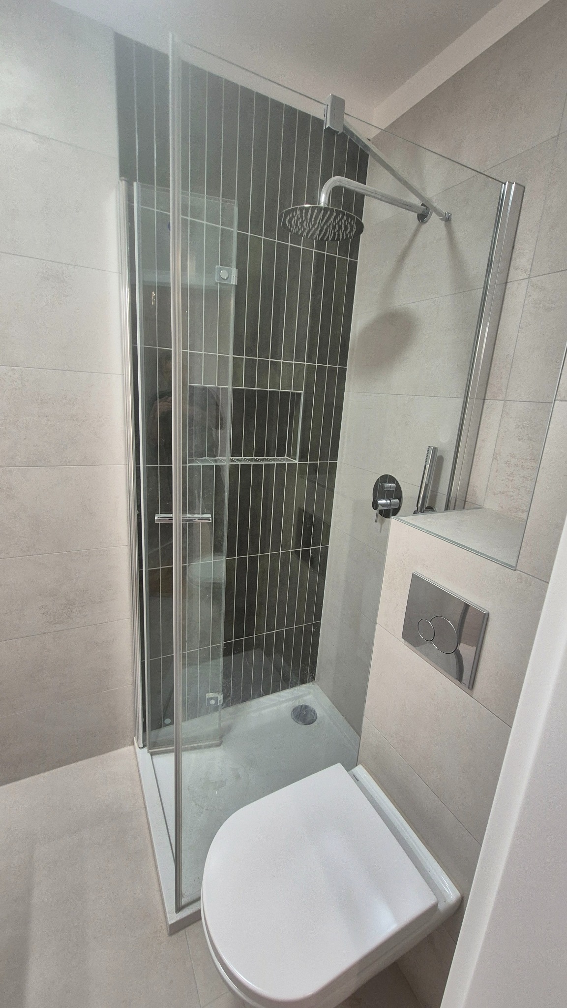 Nowoczesna łazienka po remoncie: kabina prysznicowa ze szklanymi drzwiami, podwieszana toaleta z chromowanym przyciskiem, szare płytki z ciemniejszym akcentem pod prysznicem.