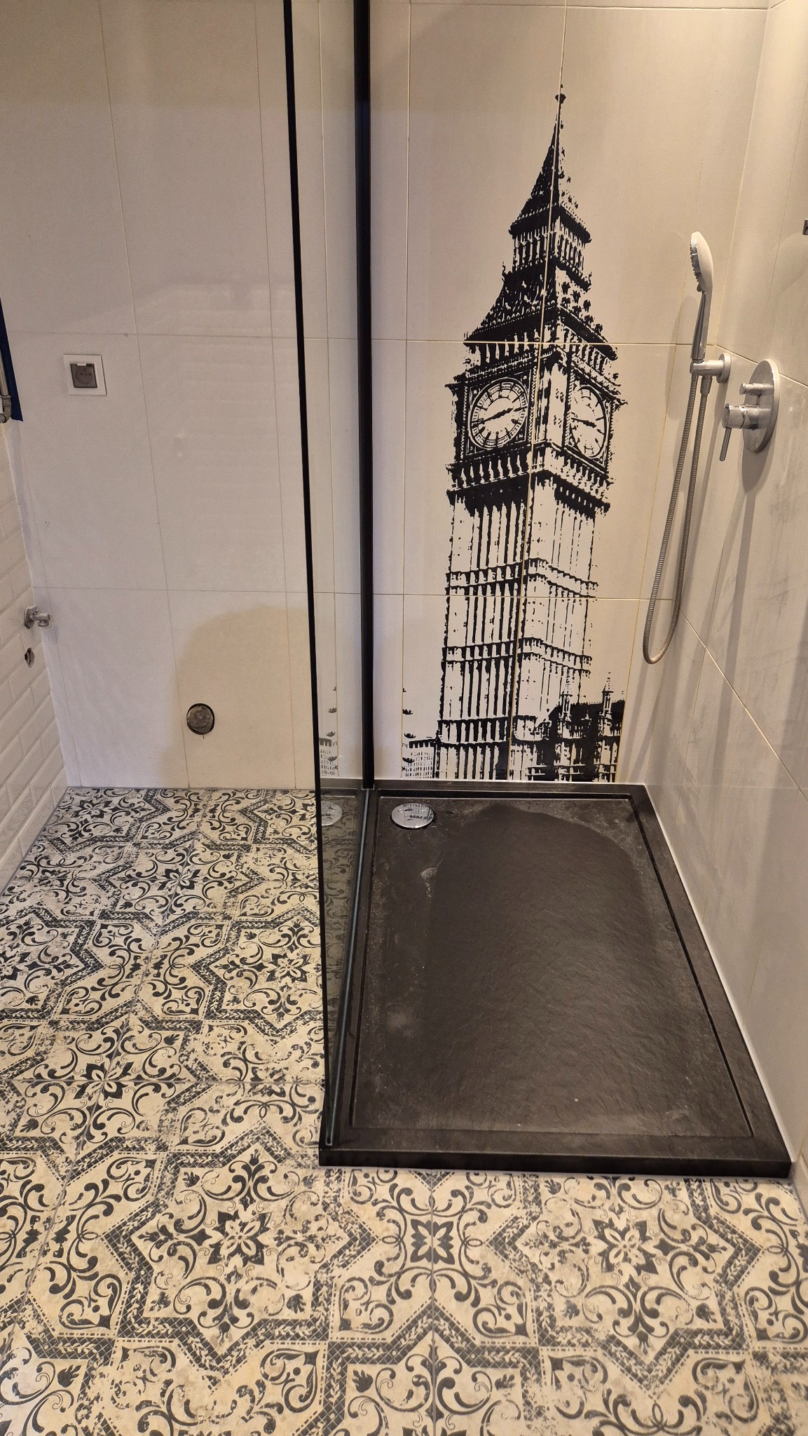 Nowoczesna kabina prysznicowa z czarnym brodzikiem i szklaną ścianką. Na ścianie dekoracja z motywem Big Bena. Podłoga wyłożona płytkami w stylu vintage.