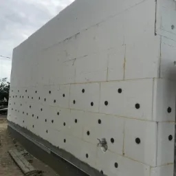 Ściana budynku w trakcie ocieplania styropianem, z widocznymi czarnymi kołkami mocującymi i otworem na instalację, na tle pochmurnego nieba.