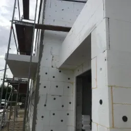 Ocieplanie budynku styropianem, widoczne rusztowanie, szare niebo w tle.