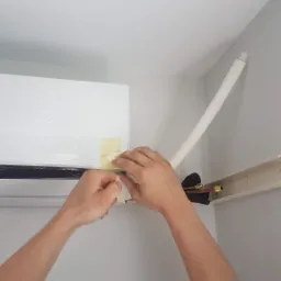 Instalacja klimatyzacji: ręce przyklejają taśmą ochronną folię do białej jednostki wewnętrznej na ścianie, widoczne rury i przewody chłodnicze.